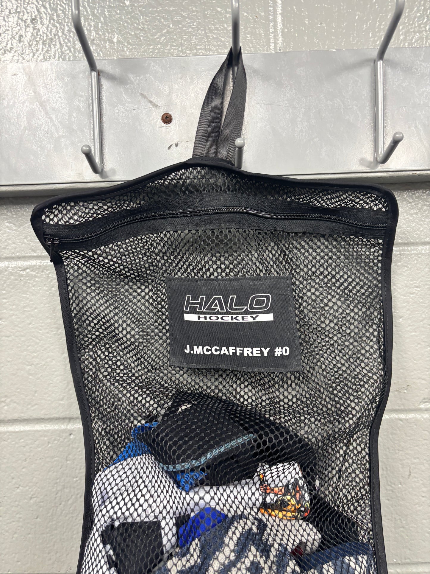 Closeup view of black Halo Hockey Stinky Boy laundry bag with custom name and number shown half full of hockey laundry hung on a locker room wall hook