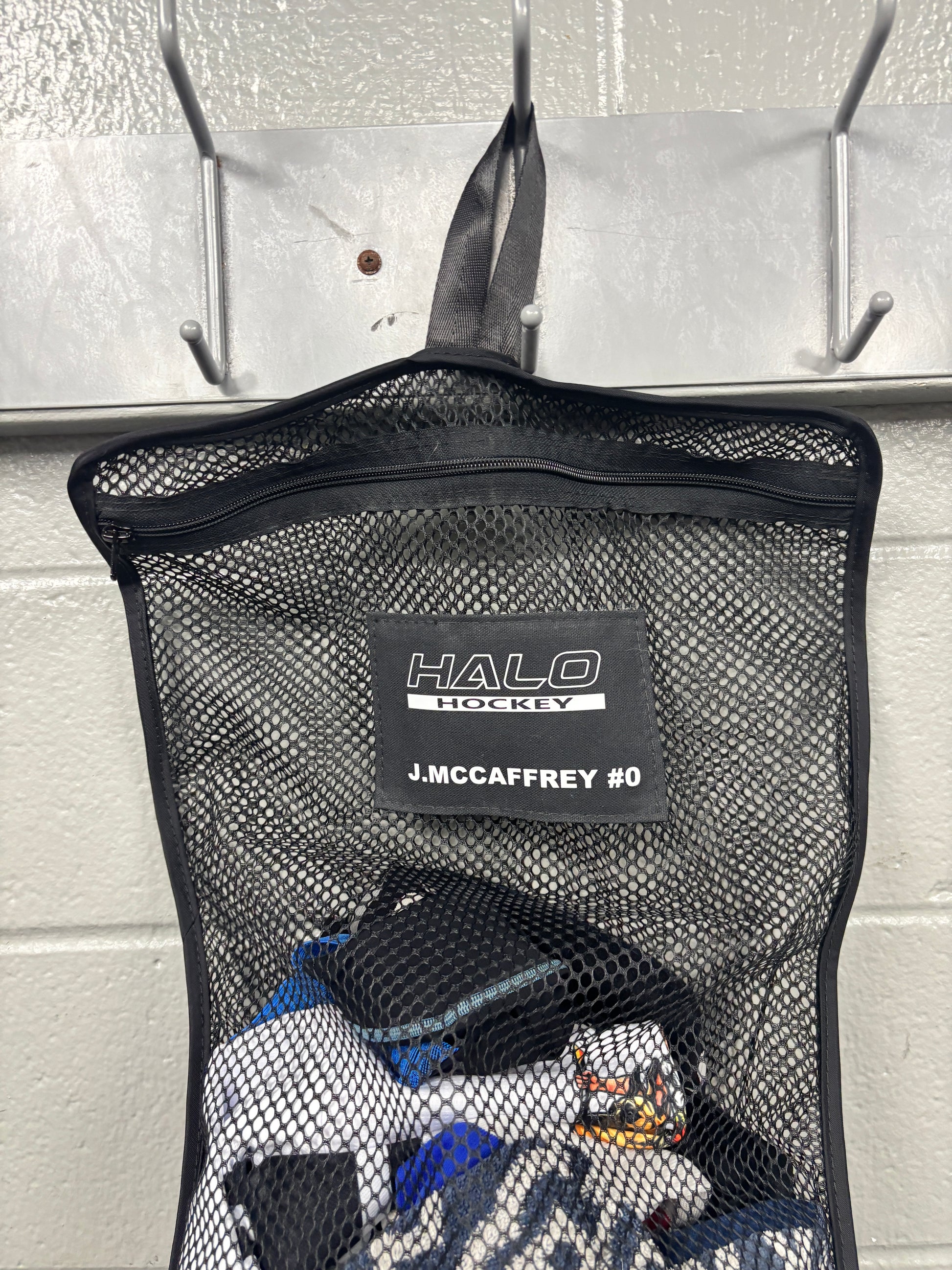 Closeup view of black Halo Hockey Stinky Boy laundry bag with custom name and number shown half full of hockey laundry hung on a locker room wall hook